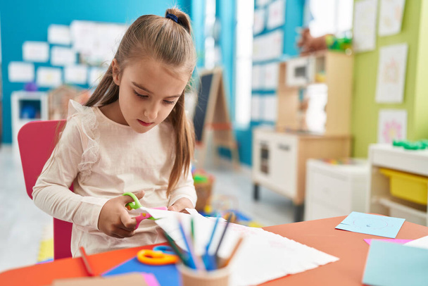 Adorable hispanic girl student sitting on table cutting paper at kindergarten - Photo, image