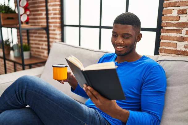 Joven afroamericano hombre leyendo libro y beber café sentado en el sofá en casa - Foto, imagen