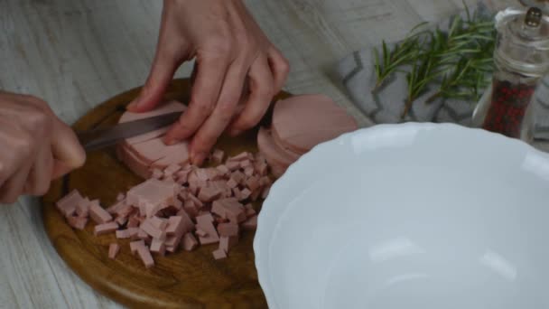 Sausage cut into circles is cut into small pieces on cutting board with knife. Female hands finely chopping sausage with knife on wooden cutting board on gray kitchen table. Preparation of meat salad. - Video