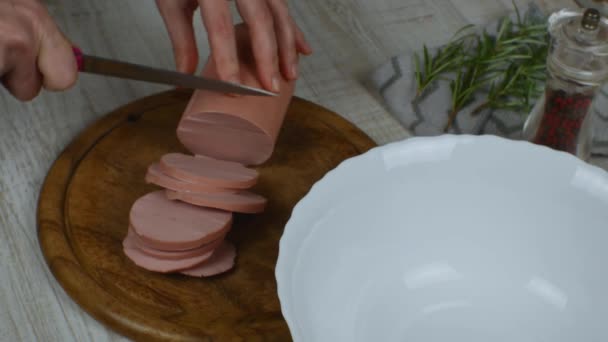 Sausage is cut on wooden cutting board with kitchen knife. Female hands slicing boiled sausage with a knife on a wooden cutting board on a gray kitchen table. Pepper grinder, rosemary sprig. Close-up. - Materiał filmowy, wideo