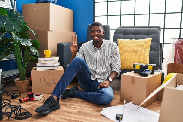 African american man sitting on the floor at new home smiling with happy face winking at the camera doing victory sign with fingers. number two.  - Foto, afbeelding