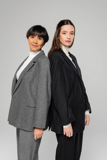 asian mother and daughter in stylish suits looking at camera while standing back to back isolated on grey - Foto, Imagem