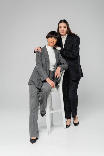 full length of young asian woman in black suit embracing stylish mother posing on white chair on grey background - Φωτογραφία, εικόνα