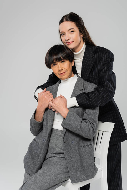 brünette asiatische Frau umarmt stilvolle Mutter sitzt auf Stuhl isoliert auf grau - Foto, Bild