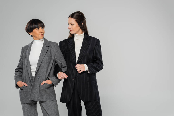 asian mother and daughter wearing stylish suits and looking at each other while standing isolated on grey - Фото, изображение