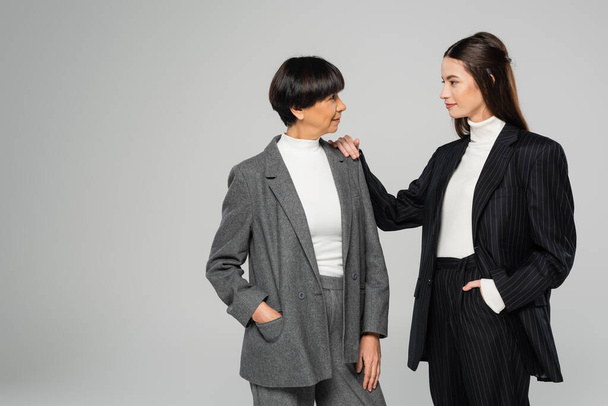 brunette woman in black suit touching shoulder of elegant asian mother isolated on grey - Photo, Image