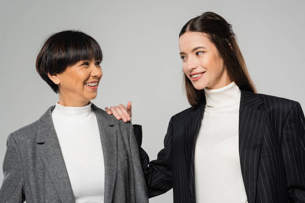 cheerful asian mother and daughter in blazers and white turtlenecks looking at each other isolated on grey - Fotografie, Obrázek
