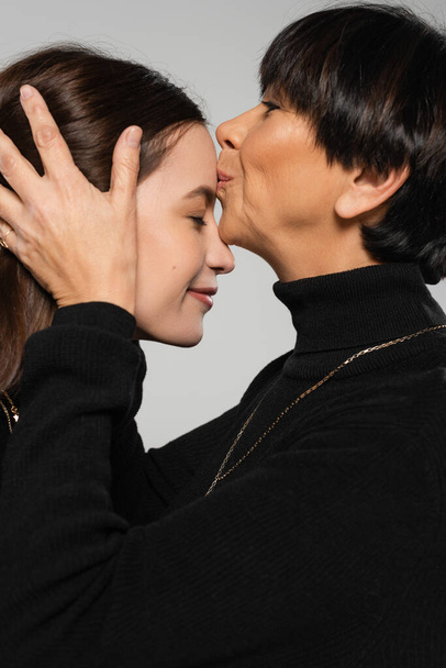 side view of middle aged asian woman in black turtleneck kissing forehead of young daughter isolated on grey - Fotó, kép