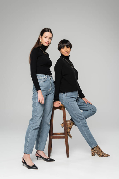 full length of asian mother and daughter in blue jeans and black turtlenecks posing near wooden stool on grey background - 写真・画像