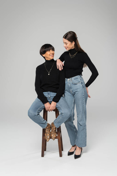 young woman in black turtleneck and jeans smiling at trendy asian mother sitting on wooden stool on grey background - Фото, изображение