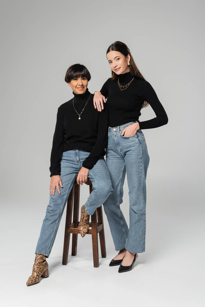 полная длина стильной женщины, стоящей с рукой в кармане джинсов возле счастливой азиатской матери, сидящей на деревянном стуле на сером фоне - Фото, изображение