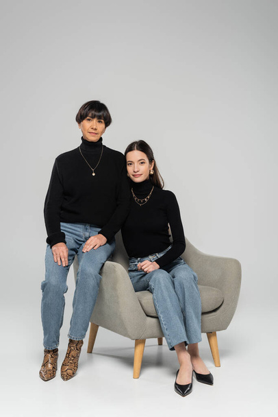 повна довжина азіатської матері і дочки в чорних черепах і джинсах позує на кріслі на сірому фоні
 - Фото, зображення