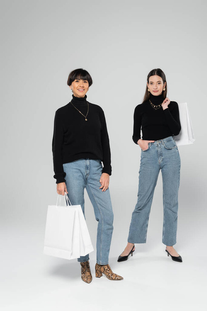 full length of asian mother and daughter in trendy outfit posing with white shopping bags on grey background - Fotografie, Obrázek
