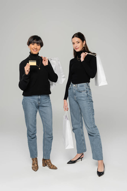 sonriente mujer asiática sosteniendo tarjeta de crédito cerca elegante hija con bolsas de compras en gris fondo - Foto, Imagen