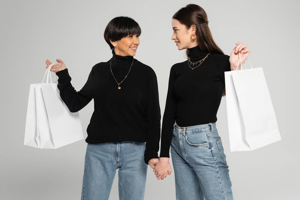 fashionable asian mother and daughter holding hands and smiling at each other while holding shopping bags isolated on grey - Фото, изображение