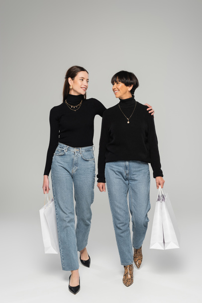 full length of stylish asian mother and daughter smiling at each other while walking with shopping bags on grey background - Foto, Imagem