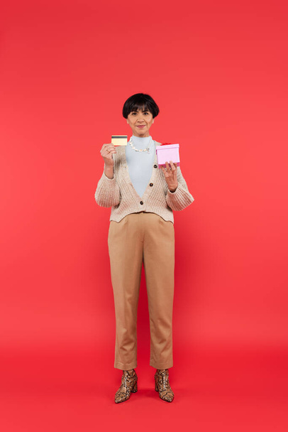 full length of happy asian woman in knitted cardigan and beige pants posing with credit card and gift box on coral background - Fotoğraf, Görsel
