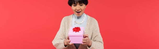 mujer asiática con estilo en cuentas de concha mirando caja de regalo con lazo rojo aislado en coral, bandera - Foto, Imagen