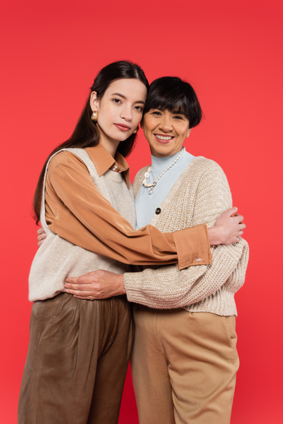 asian mother and daughter in stylish casual outfit embracing and looking at camera isolated on coral - Фото, зображення