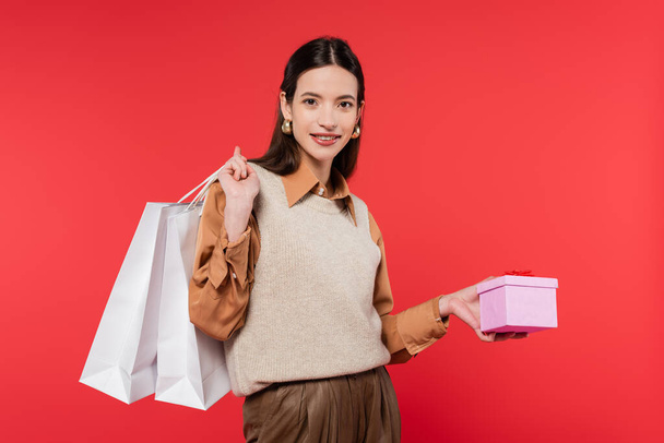young woman in stylish casual attire holding present and shopping bags while smiling at camera isolated on coral - Fotografie, Obrázek