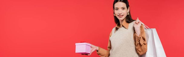 cheerful and stylish asian woman holding gift box and shopping bags isolated on coral, banner - Zdjęcie, obraz