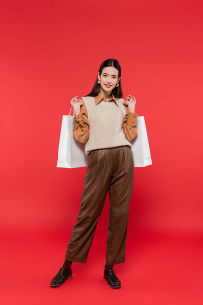 full length of smiling woman in brown pants and knitted vest standing with white shopping bags on coral background - Photo, image