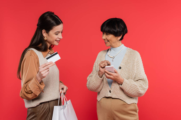 smiling asian woman pointing at smartphone near young daughter holding shopping bags and credit card isolated on coral - Foto, afbeelding