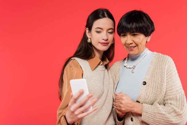cheerful asian mother and daughter looking at smartphone isolated on coral  - Foto, Bild