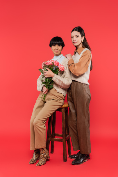 full length of young asian daughter hugging happy mother sitting on high chair and holding bouquet of flowers on coral  - Fotografie, Obrázek