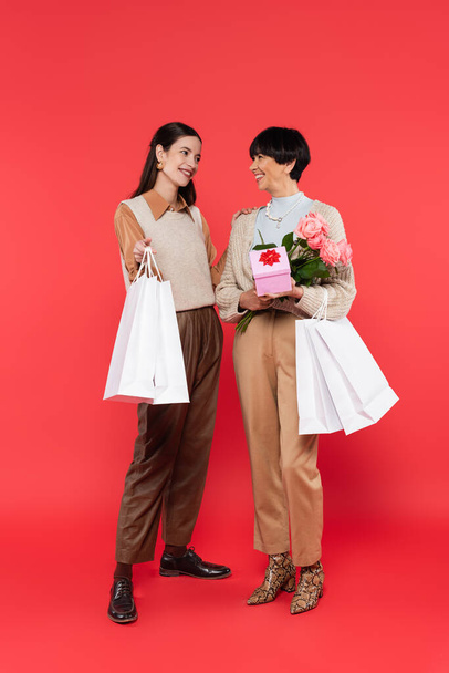 full length of young asian daughter with shopping bags looking at happy mother holding flowers and gift box on coral - Photo, Image
