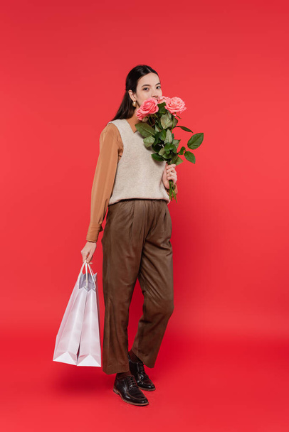 full length of young asian woman holding roses and shopping bags on coral background  - Photo, image
