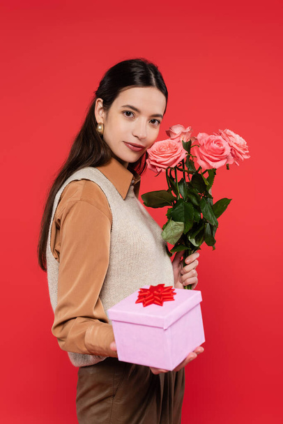 νεαρή γυναίκα από την Ασία κρατώντας μπουκέτο τριαντάφυλλα και κουτί δώρου απομονώνονται σε κοράλλια  - Φωτογραφία, εικόνα