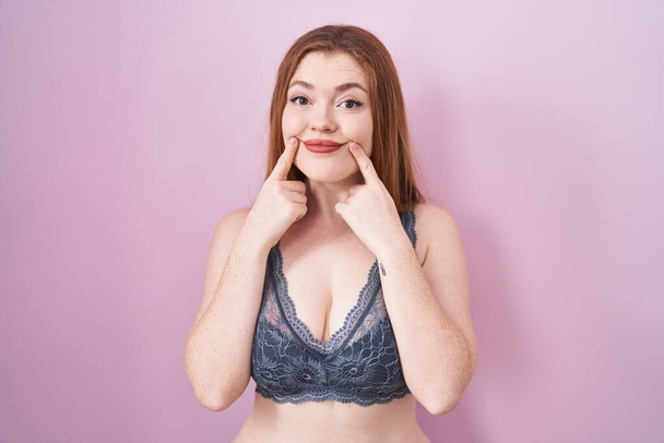 Redhead woman wearing lingerie over pink background smiling with open mouth, fingers pointing and forcing cheerful smile  - Fotó, kép