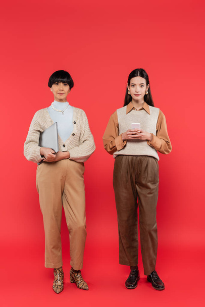 full length of asian mother and daughter in stylish clothes holding devices on coral  - Photo, Image