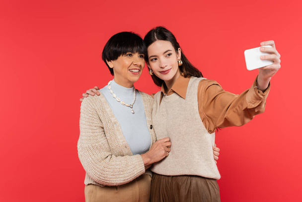 young asian woman in knitted vest taking selfie with happy mother isolated on coral  - Photo, image