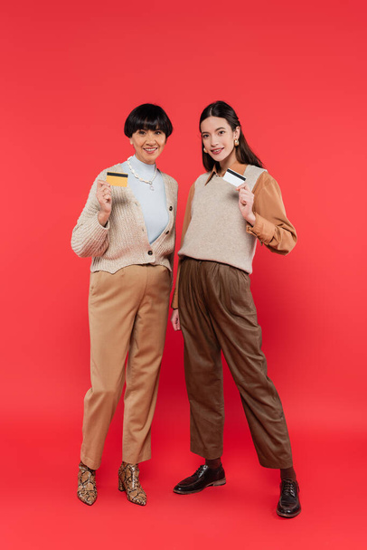 full length of happy asian mother and daughter in stylish clothes holding credit cards on coral background  - Foto, Imagem