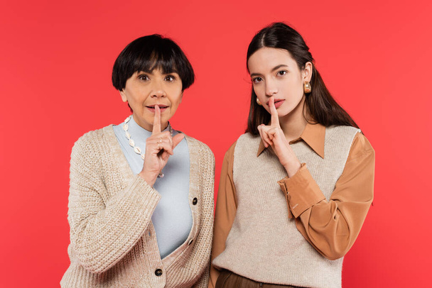 brunette asian mother and daughter in stylish clothes showing hush sign isolated on coral  - Photo, Image