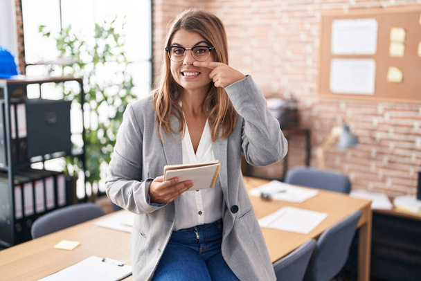 Νεαρή Ισπανίδα που εργάζεται στο γραφείο φορώντας γυαλιά δείχνοντας με το χέρι το πρόσωπο και τη μύτη, χαμογελώντας χαρούμενη. έννοια ομορφιά  - Φωτογραφία, εικόνα