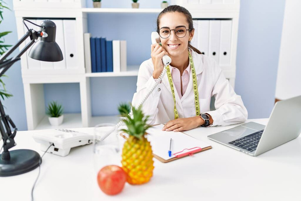 Young hispanic woman wearing nutritionist uniform talking on the telephone at clinic - Photo, Image