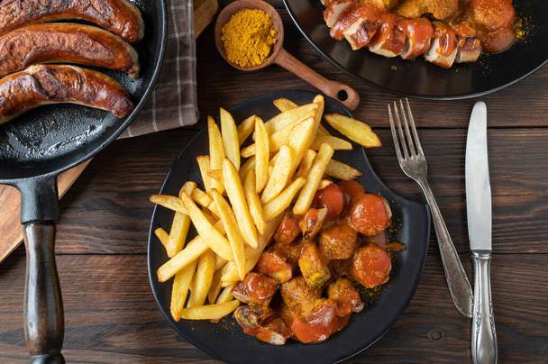 Traditional german fast food meal with currywurst or curry sausage with french fries and delicious curry sauce. Served on plates with cutlery on wooden and rustic table background. Flat lay - Photo, Image