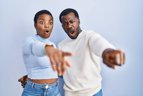 Junges afrikanisch-amerikanisches Paar steht vor blauem Hintergrund und zeigt mit dem Finger überrascht nach vorne, offener Mund erstaunter Ausdruck, etwas auf der Vorderseite  - Foto, Bild