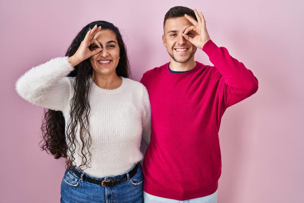 Young hispanic couple standing over pink background doing ok gesture with hand smiling, eye looking through fingers with happy face.  - Фото, зображення