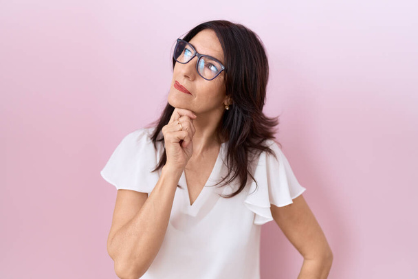 Middle age hispanic woman wearing casual white t shirt and glasses with hand on chin thinking about question, pensive expression. smiling with thoughtful face. doubt concept.  - Photo, image