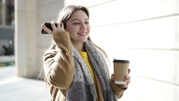 Νεαρή ξανθιά γυναίκα ακούει ηχητικό μήνυμα από το smartphone πίνοντας καφέ στο δρόμο - Φωτογραφία, εικόνα