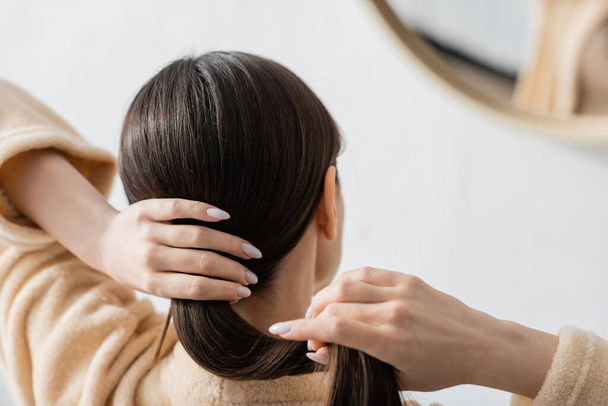 πίσω όψη της νεαρής μελαχρινής γυναίκας προσαρμογή λαμπερά μαλλιά κοντά στον καθρέφτη στο μπάνιο - Φωτογραφία, εικόνα