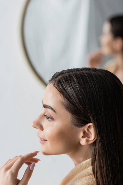 πλαϊνή άποψη του χαμογελαστού νεαρή γυναίκα κοιτάζοντας μακριά κοντά θολή καθρέφτη στο μπάνιο  - Φωτογραφία, εικόνα