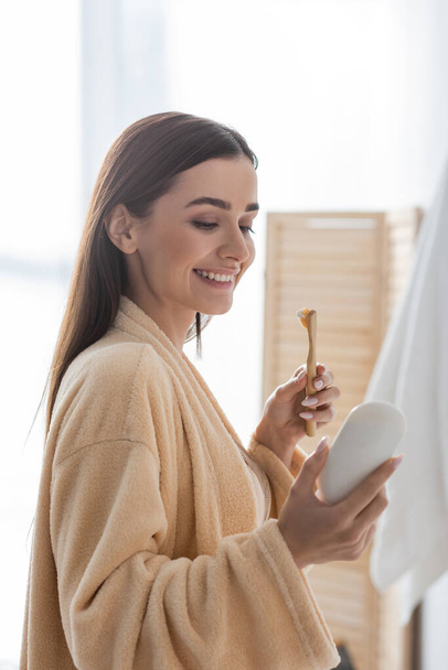 счастливая молодая женщина, держащая зубную щетку и глядя на тюбик с зубной пастой утром  - Фото, изображение