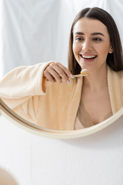 reflection of cheerful young woman with toothbrush in bathroom mirror  - Fotó, kép