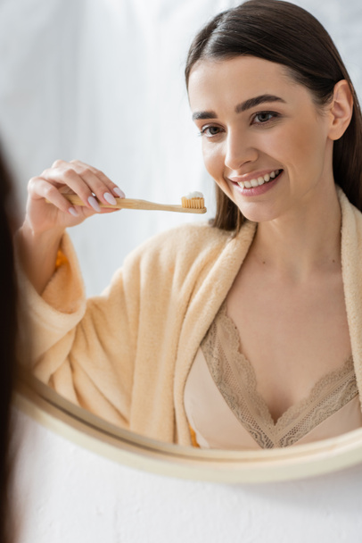 giovane e allegra donna che tiene lo spazzolino da denti e guarda lo specchio in bagno - Foto, immagini