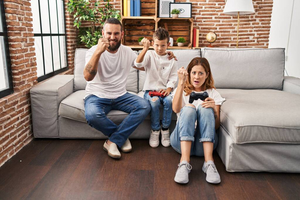 Οικογένεια τριών παιδιών που παίζουν βιντεοπαιχνίδι καθισμένοι στον καναπέ ενοχλημένοι και απογοητευμένοι φωνάζοντας από θυμό, φωνάζοντας τρελοί από θυμό και σηκώνοντας το χέρι  - Φωτογραφία, εικόνα